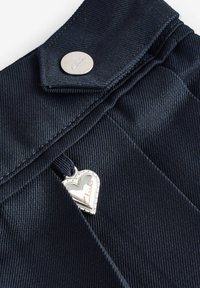 Il tessuto denim blu scuro presenta un bottone a pressione in metallo e un ciondolo a forma di cuore argentato con la scritta "Clarks" incisa. Superficie texturizzata visibile.