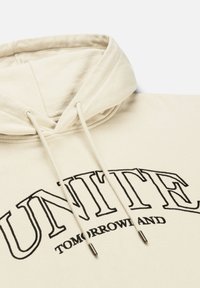 Beige hoodie met trekkoorden, met geborduurde zwarte tekst "UNITE TOMORROWLAND." Zachte stof met een relaxte fit en een voorzak in kangoeroestijl.