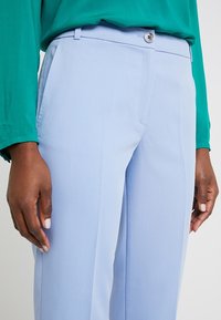 Jasnoniebieskie spodnie wykonane z gładkiego materiału, z płaskim przodem, pojedynczym zapięciem na guzik oraz bocznymi kieszeniami, w połączeniu z zieloną bluzką.