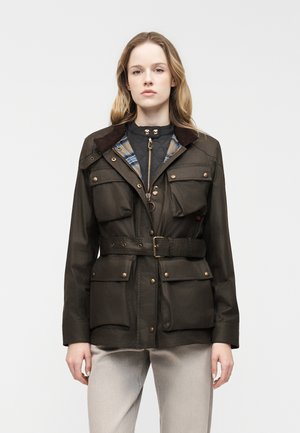 Ung kvinde iført en olivengrøn bæltejakke med flere forlommer over en quiltet vest og lyse bukser, stående oprejst.