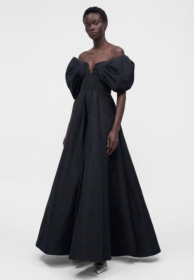 Γυναίκα που φορά μακρύ μαύρο φόρεμα χωρίς ώμους με μεγάλα φουσκωτά μανίκια και πλισέ λεπτομέρειες, στέκεται μπροστά σε μια απλή φόντο.