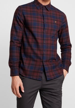 Uomo che indossa una camicia a maniche lunghe con motivo a quadri rosso e blu navy e pantaloni grigio scuro, con una mano in tasca.