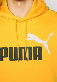 Personne portant un sweat à capuche jaune avec le texte Puma en noir et un logo puma blanc sur la poitrine, visible du cou jusqu'au milieu du torse.