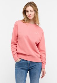 Felpa rosa con maniche lunghe, polsini a coste e scollo rotondo. Presenta un piccolo logo sul petto e una vestibilità comoda. Abbinata a jeans blu.