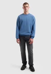 Jongeman staat rechtop, gekleed in een blauwe sweatshirt met ronde hals, zwarte spijkerbroek en zwarte veterschoenen tegen een eenvoudige lichte achtergrond.