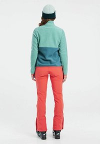 Polarowy szal w jasnym zielonym i ciemnym niebieskim kolorze, z wysokim kołnierzem; zestawiony z jaskrawymi pomarańczowymi spodniami narciarskimi i czarnymi butami narciarskimi, z zamkami bocznymi.