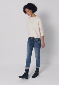Femme aux cheveux bouclés portant un pull léger, un jean bleu retroussé et des bottines noires, debout devant un fond clair uni.