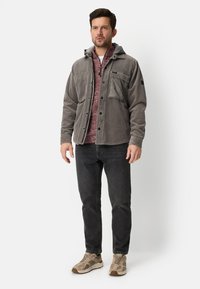 Grijze corduroy jas met een capuchon, voorzien van een voorzak met rits, gedragen over een rode, gestructureerde trui, gecombineerd met zwarte spijkerbroek en beige sneakers.