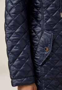 Abrigo acolchado en color navy con una textura brillante, costuras diagonales y un bolsillo con solapa adornado con un botón decorativo en el lado izquierdo.