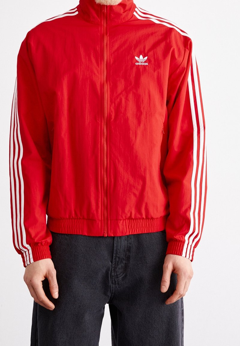 Röd Adidas-jacka med vit tre-stripesdetaljering på ärmarna, hög krage, elastisk midja och mjukt, lätt tyg.