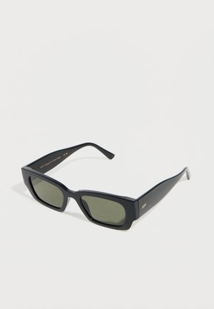 Schwarze rechteckige Sonnenbrille mit dicken Rahmen und dunklen Gläsern, die auf einer weißen Oberfläche liegt und ein kleines goldenes Logo am Bügel zeigt.