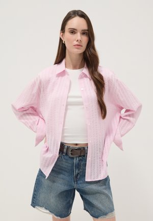 Vrouw met lang bruin haar draagt een lichtroze bloes met knopen over een witte cropped top en spijkershorts met een zwarte riem, staand met handen in de zij.
