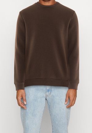 Sudadera marrón con cuello redondo, puños acanalados y cintura acanalada. Llevada sobre unos vaqueros azul claro, mostrando una textura de tela suave.