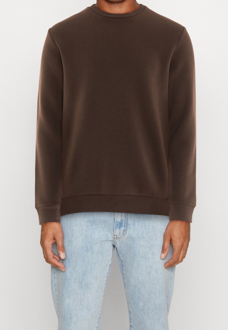Sudadera marrón con cuello redondo, puños acanalados y cintura acanalada. Llevada sobre unos vaqueros azul claro, mostrando una textura de tela suave.