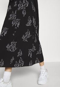 Jupe noire avec des figures abstraites en lignes blanches associée à des chaussettes blanches et des baskets blanches épaisses sur une personne debout.