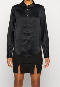 Camicia nera di satin a maniche lunghe con abbottonatura frontale, colletto e orlo curvo, abbinata a una gonna nera con due spacchi laterali.