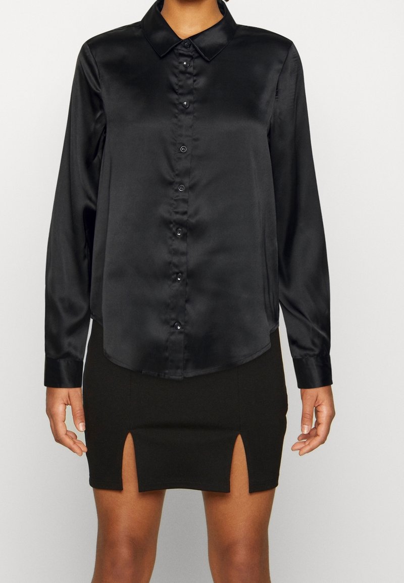 Camicia nera di satin a maniche lunghe con abbottonatura frontale, colletto e orlo curvo, abbinata a una gonna nera con due spacchi laterali.
