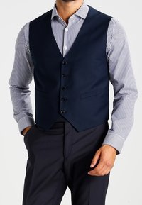 Mann, der eine marineblaue Weste über einem langärmeligen, weiß-blauen gestreiften Hemd und einer schwarzen Anzughose trägt.