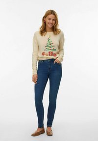 Pull crème avec un motif de sapin de Noël, agrémenté de cadeaux, associé à un jean bleu ajusté et des chaussures plates marron. Look décontracté.