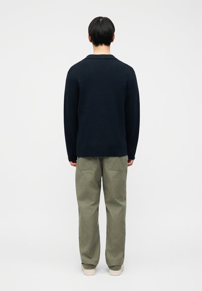 Homme debout de dos portant un haut à manches longues noir, un pantalon vert olive et des baskets blanc cassé sur un fond uni.