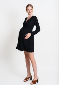 Abito premaman nero con scollo a V, maniche lunghe e vita annodata. Sandali con tacco marroni completano il look. Tessuto liscio e vestibilità sartoriale.