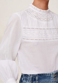 Blouse blanche à col à volants, présentant des rayures verticales et un détail en dentelle sur le corsage, associée à un short en denim taille haute.