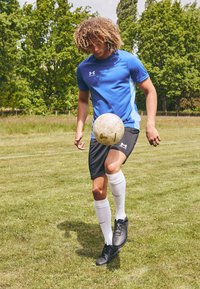 Blauses, kurzärmliges Sportshirt mit schwarzen Shorts, weißen kniehohen Socken und schwarzen Fussballschuhen. Ein Spieler balanciert einen beigen Fussball.