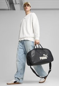 Sac de sport en cuir noir avec logo "PUMA" blanc, design structuré, doubles poignées, sangle ajustable, et texture lisse.
