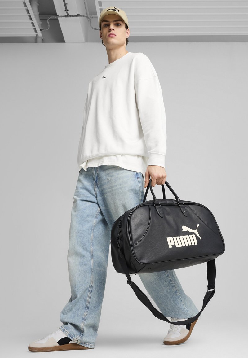 Sac de sport en cuir noir avec logo "PUMA" blanc, design structuré, doubles poignées, sangle ajustable, et texture lisse.