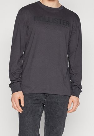 Langærmet, mørkegrå bomulds T-shirt med "HOLLISTER" logo i sort på brystet og blød tekstur. Båret med mørke jeans.