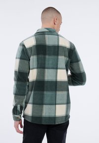 Man die met zijn rug naar de camera staat, gekleed in een groen en wit groot geruit flanellen shirt met zwarte broek, tegen een simpele witte achtergrond.