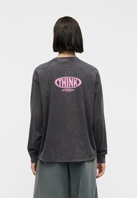 Άτομο με κοντά σκούρα μαλλιά που φοράει φθαρμένο μαύρο φούτερ με ροζ κείμενο "THINK DIFFERENT" στην πλάτη, συνδυασμένο με φαρδιά πράσινα παντελόνια.