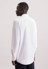 Camicia bianca a maniche lunghe realizzata in tessuto morbido, con colletto classico, polsini con un bottone e orlo curvo; design semplice senza motivi.