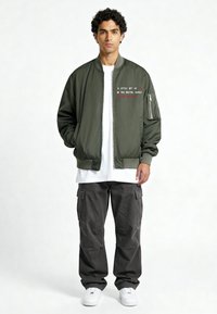 Legergroene bomberjack met een ritssluiting aan de voorkant, geribbelde manchetten, een wit t-shirt eronder, cargobroek en witte sneakers. Geborduurde tekst op het jack.