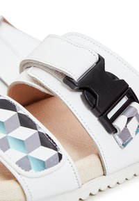 Hvide sandaler med en sort spænde, der har et geometrisk mønster i grå og teal. Blød tekstureret inderside i en neutral farve.