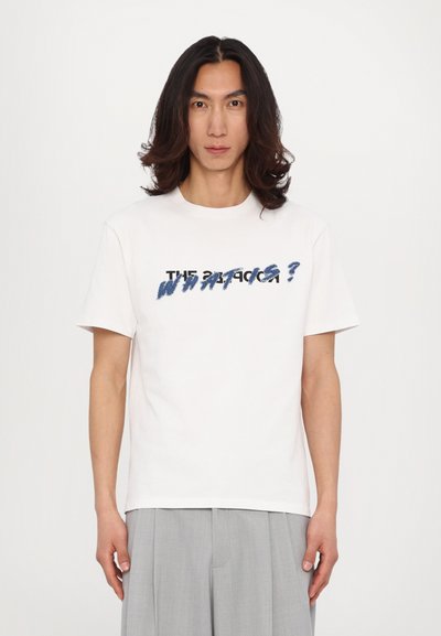 Witte katoenen t-shirt met korte mouwen, met tekst in zwart en blauw. De tekst bevat "DE" en "WAT IS?" in een gedurfde, grafische stijl.
