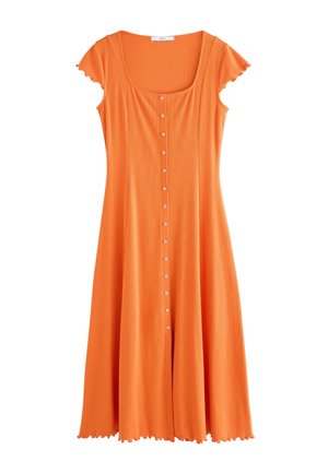 Žena stojí na pláži v plynulých oranžových maxi šatách na gombíky a čiernych žabkách s vlnami oceánu v pozadí.