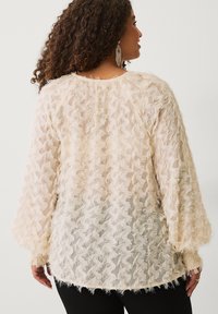 Vrouw met krullend haar, gekleed in een crèmekleurige, gestructureerde longsleeve top en zwarte broek, met de rug naar de camera gedraaid, waarbij details van de achterkant en hangende oorbellen zichtbaar zijn.