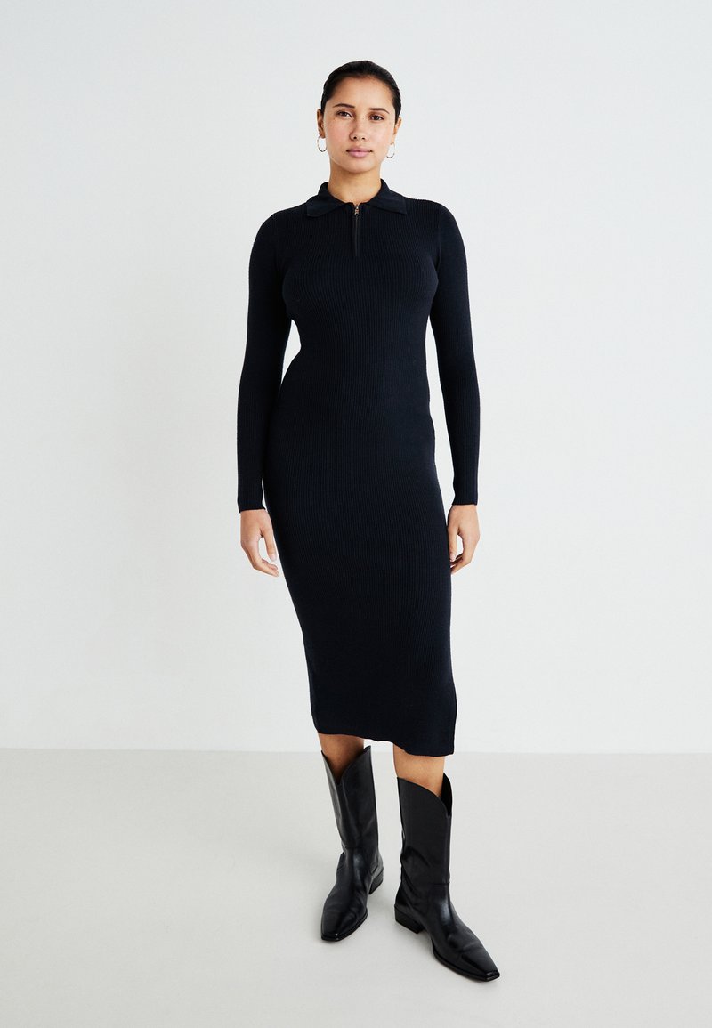 Black, ribbed knit dress with long sleeves and a zippered collar. Paired with pointed-toe black boots, featuring a high shaft.