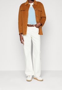 Osoba w brązowej kurtce zapinanej na guziki, jasnoniebieskim swetrze w sploty, białych spodniach z brązowym paskiem i białych tenisówkach z gumowymi podeszwami.