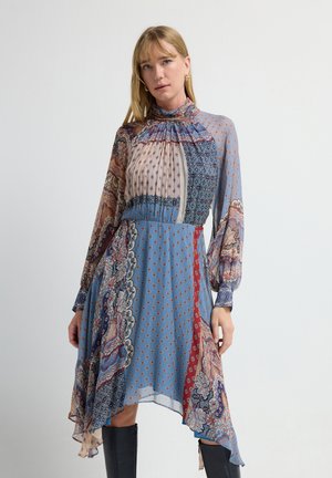 Vrouw in een hoge hals, lange mouw patchwork jurk met paisley en geometrische patronen in blauw, rood en beige, gecombineerd met zwarte laarzen tot de knie.