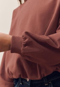 Ruskea collegepaita pussihihoilla ja resorillisilla hihoilla. Pehmeä, teksturoitu kangas rentolla istuvuudella, yhdistetty farkkuhousuihin.