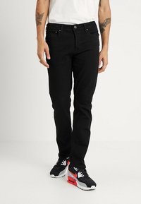Black denim jeans with a slim fit, featuring five pockets, a button closure, and subtle stitching details. Worn with black and red sneakers.