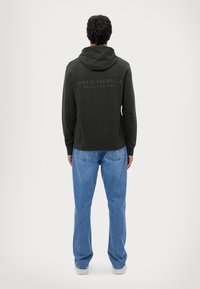 Mørkegrønn hettegenser med preget "Armani Exchange"-logo på baksiden, kombinert med lyseblå denimjeans, vist fra baksiden.