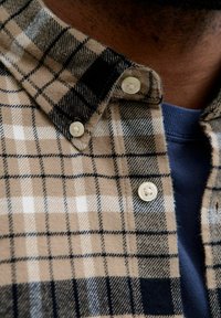 Braun und schwarze karierte Hemd mit weißen Akzenten; Button-Down-Kragen; weiche Textur; mit zwei Knöpfen; übereinander getragen über einem blauen Hemd.