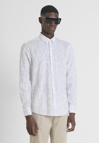 Witte button-down shirt met een subtiel gouden abstract patroon, lange mouwen en een klassieke kraag. Model draagt een oversized zwart zonnebril.