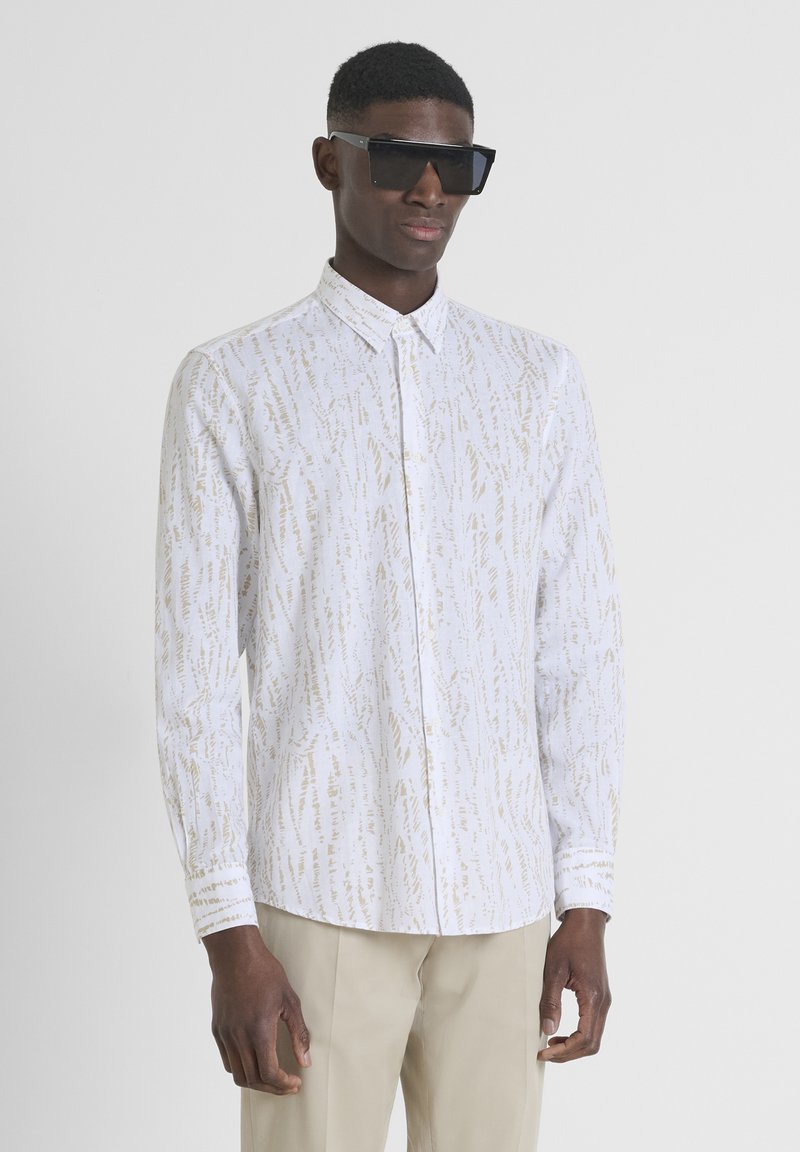 Witte button-down shirt met een subtiel gouden abstract patroon, lange mouwen en een klassieke kraag. Model draagt een oversized zwart zonnebril.