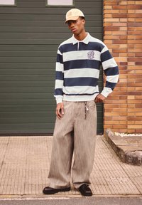 Prugasta polo majica dugih rukava u tamnoplavoj i svijetloplavoj boji s izvezenim logotipom, uparen s bež širokim hlačama i crnim cipelama.