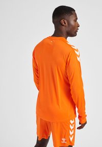 Oranje sportuniform met een lange mouw top en shorts. De top heeft witte chevron accenten op de schouders en een logo nabij de zoom.
