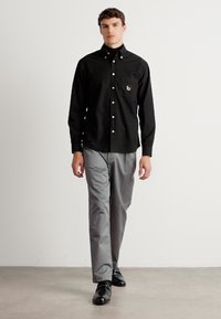 Hombre caminando con una camisa negra abotonada con bolsillo en el pecho y pantalones grises frente a un fondo blanco liso.